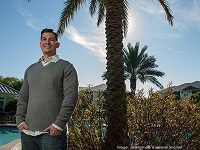 Man standing by a palm tree, bush and pool wearing gray sweater and jeans