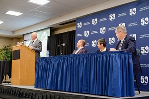 A speaker at the podium while 3 men in suits listen blue UNF logo background and table
