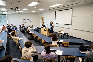 A professor lecturing to her classroom full of students next to a podium and whiteboard