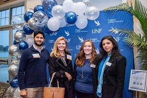 4 people smiling in business attire with a blue background with the UNF logo and balloons