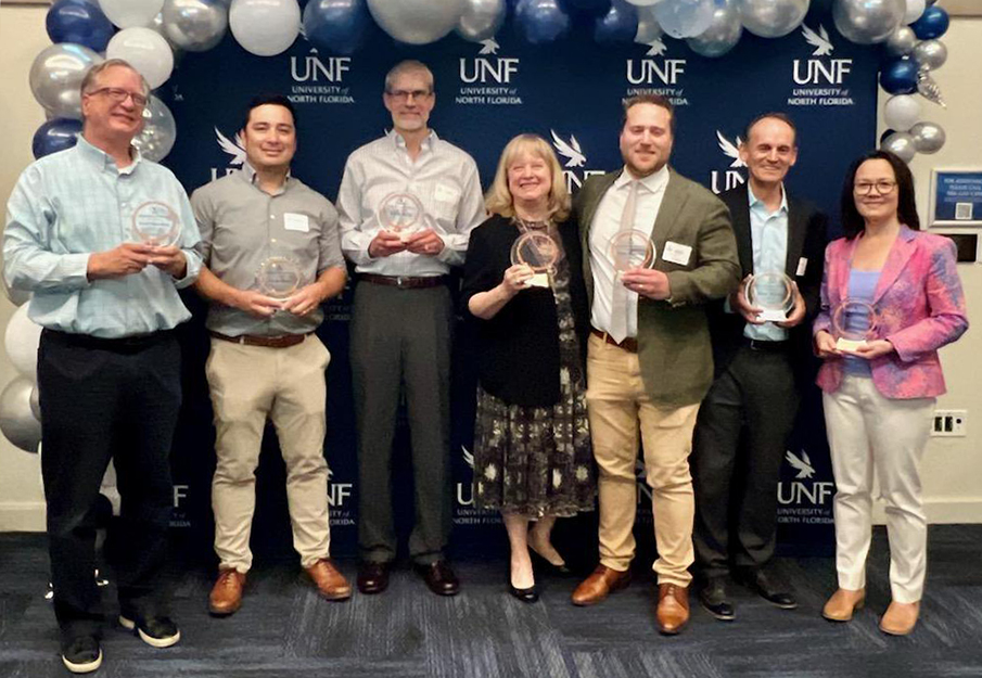 David Bauerlein, Chris Hong, Carla Miller, Nate Monroe, Mark Wood and Anna Brosche standing in front of a UNF backdrop holding their awards