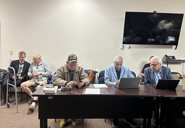 Three men at a desk working on laptops and phones while two men talk in the back