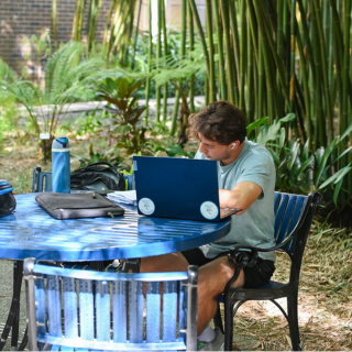 Student sitting at a blue table outdoors, writing on a notebook