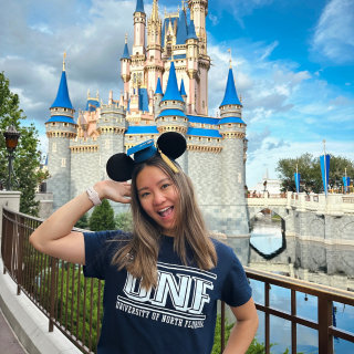 Student wearing a UNF tshirt stands in front of the Walt Disney World castle and smiles