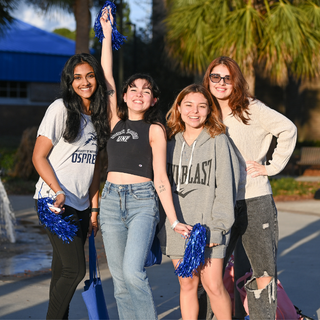Four students stand in front of osprey fountain and smile while holding pom poms.
