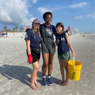 Three students stand on the sand and smile. One student is holding a yellow bucket.