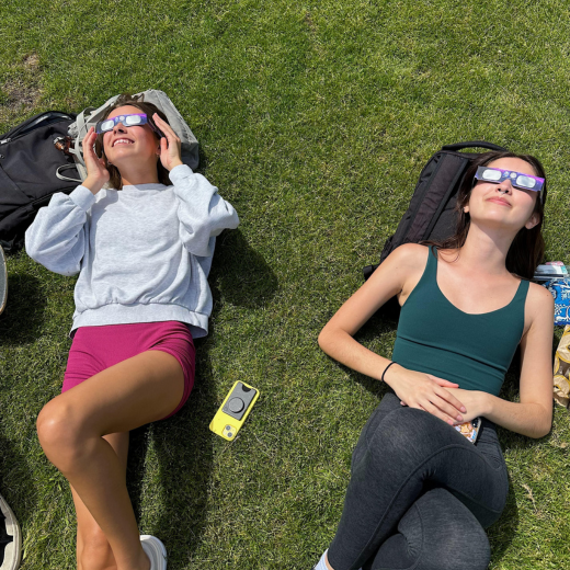 two students laying in the grass looking up while wearing eclipse glasses and smiling