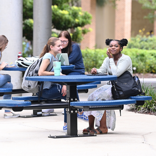 Two students sit at a blue table and talk. Students blurred in background