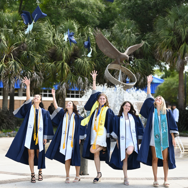 5 students stand in front of a fountain of water and throw up their graduation caps while smiling