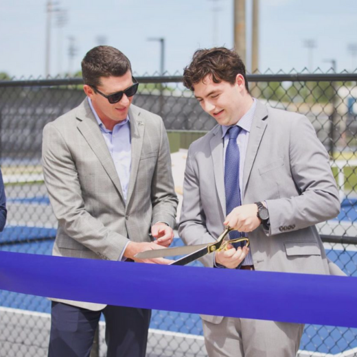 Two men stand in suits. One is cutting a blue ribbon with an oversized pair of scissors