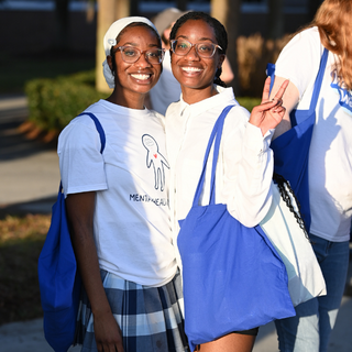 Two Students stand and smile. One student is holding up a peace sign. 