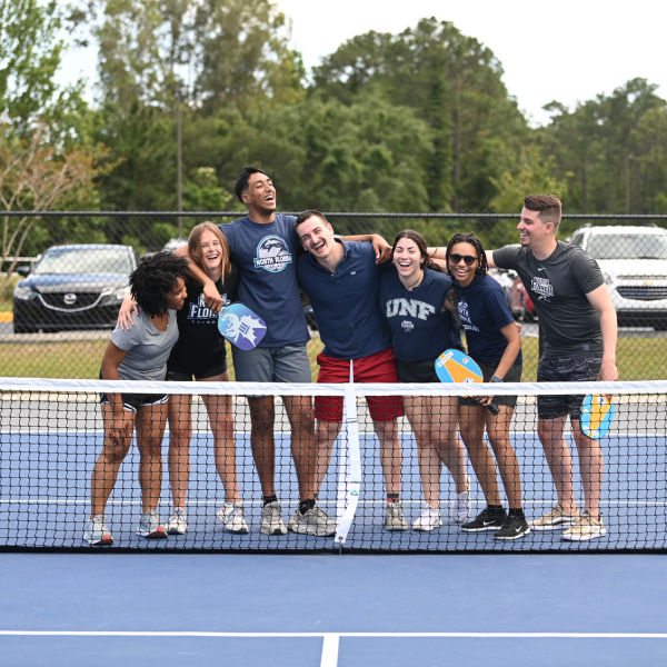 7 students standing in front of a pickleball net, holding pickleball paddles and laughing