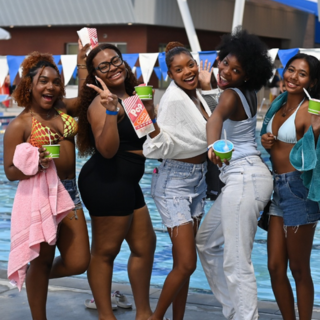 five girls stand and smile in front of a pool holding snacks.