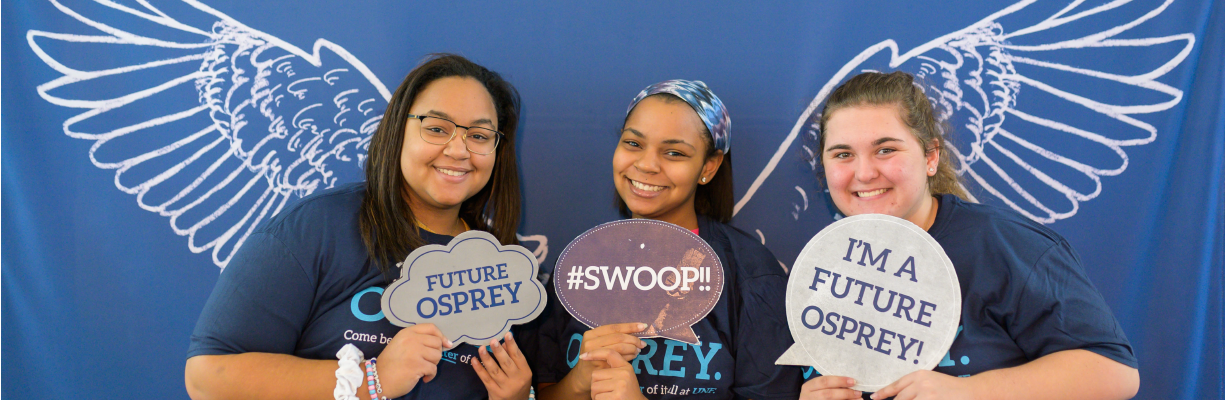Three girls smiling holding future osprey signs wearing UNF t-shirts and in front of a wings backdrop