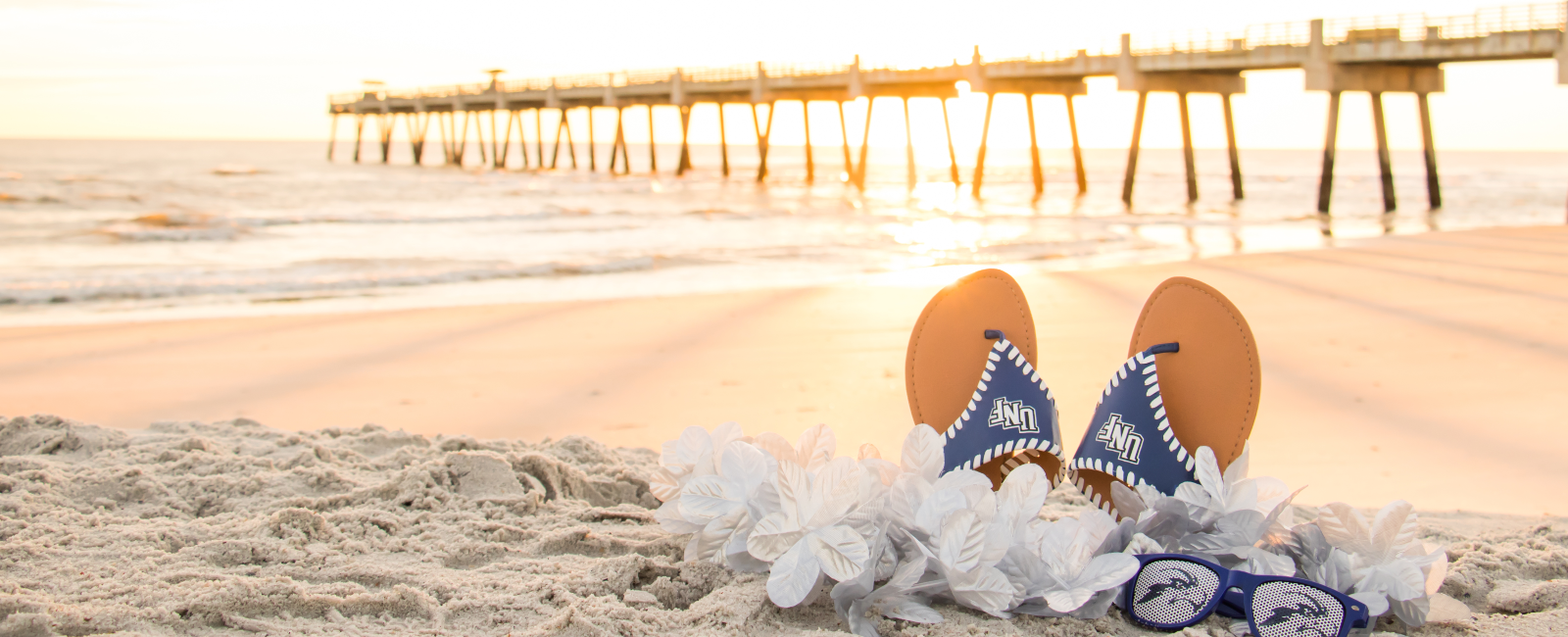 Pair of UNF flip flops, lei necklaces, and sunglasses on the beach with pier in background