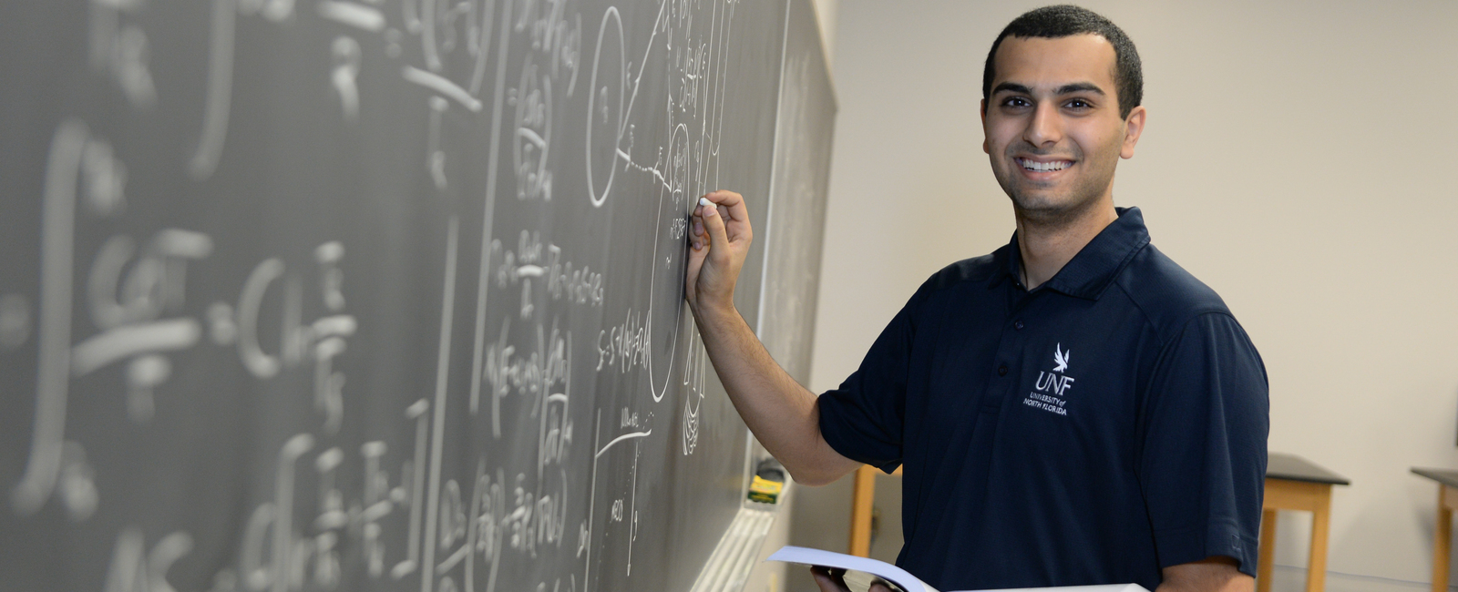 Male student in a UNF polo holding a book in front of a chalkboard with formulas written on it