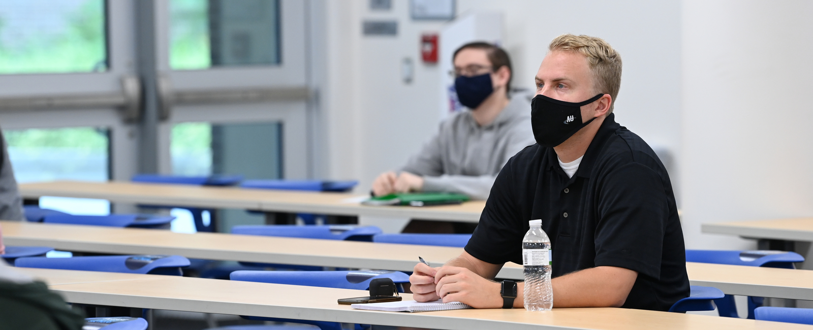 Older male student sitting in a classroom wearing a face mask and holding a pen taking notes