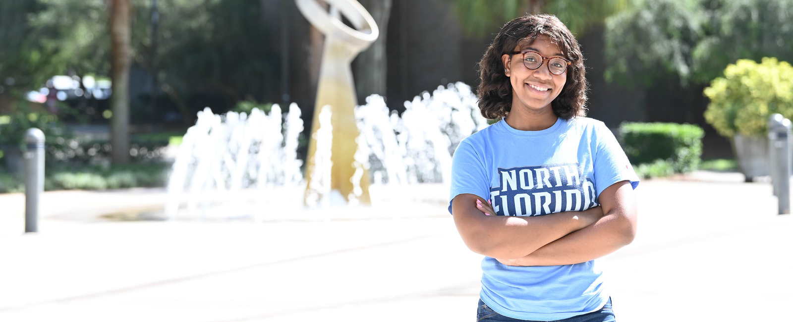 Woman in a bright blue UNF t-shirt smiling with her arms crossed and in front of UNF fountain