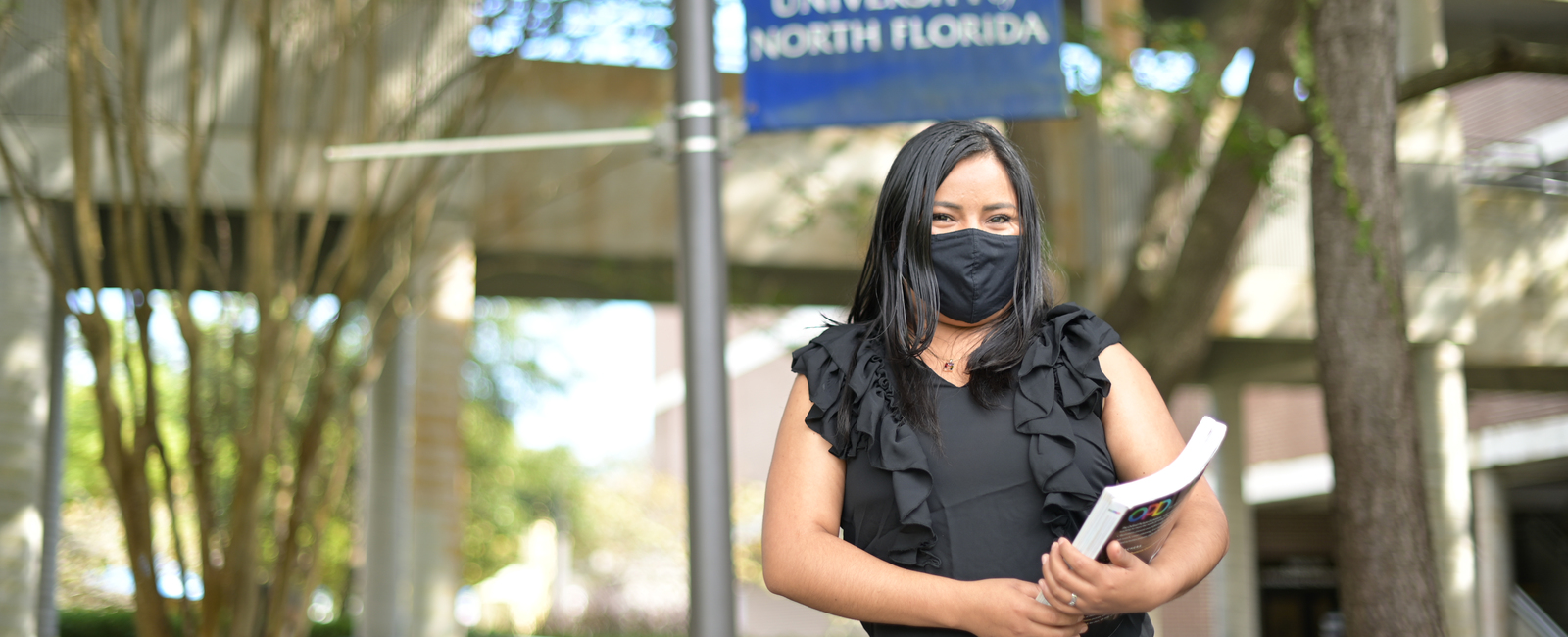 Female student holding books and wearing a face mask while standing outside on campus