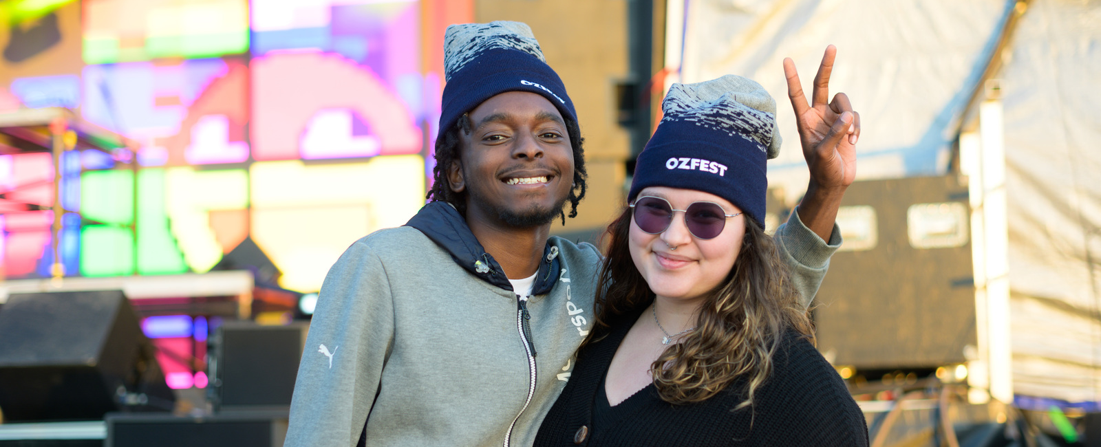 Male and female student in Oz Fest beanies with a colorful stage backdrop