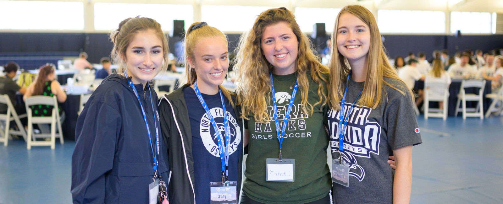 Four female students in UNF attire and lanyards inside a gymnasium smiling while at orientation