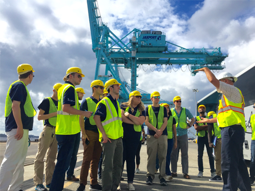 Group of students on a job site wearing yellow safety vests and hard hats with a crane in the back