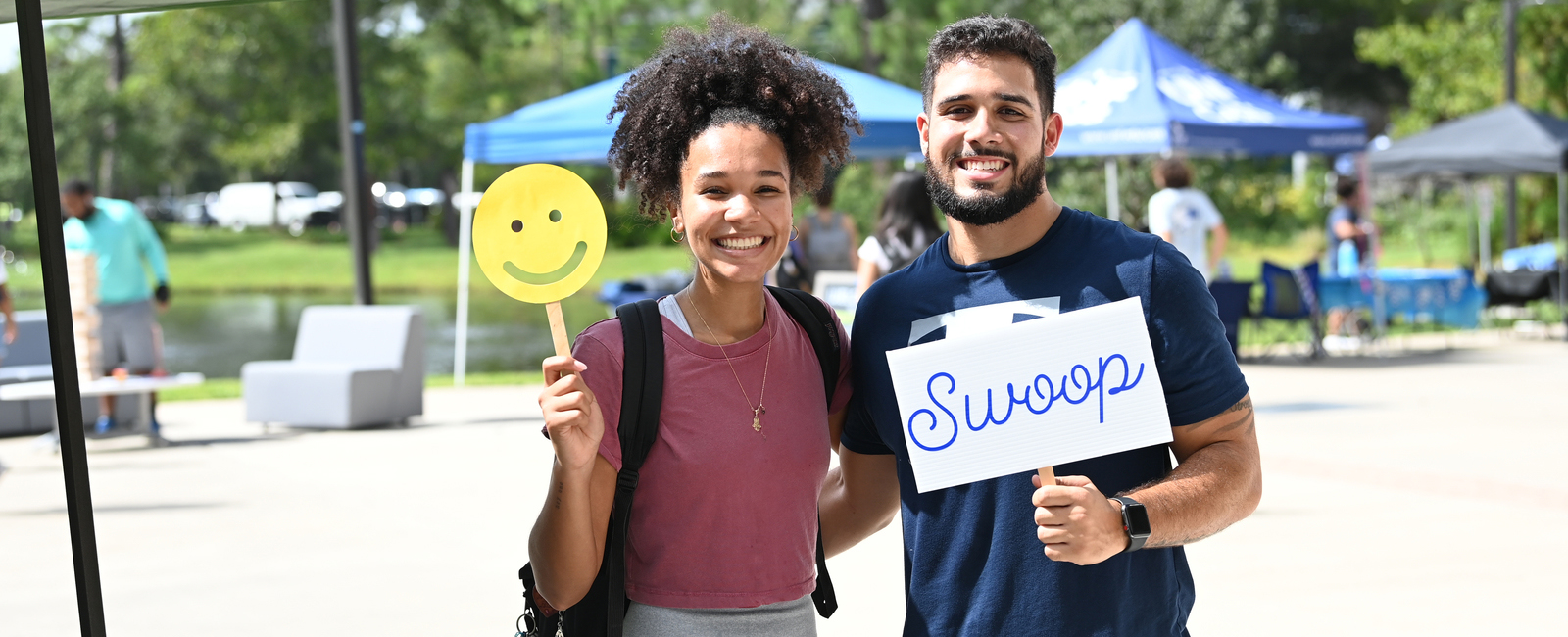 Male holding a Swoop sign and female holding a smiley face sign while both smiling outside the student union