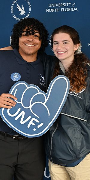 two students in front of a UNF backdrop with one holding a UNF foam finger