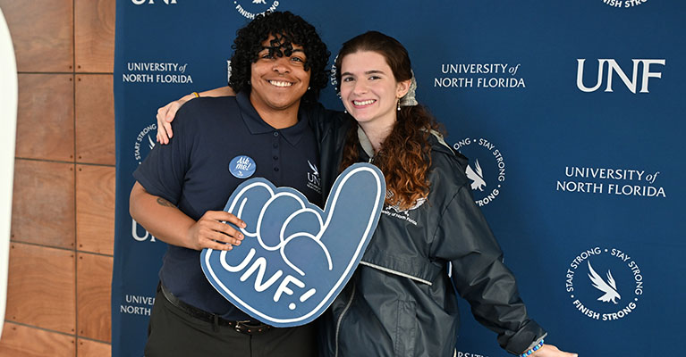 Students smiling in front of a UNF backdrop while holding a UNF foam finger