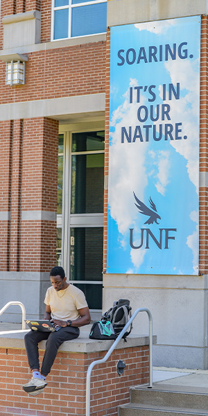 A student working on a laptop in front of a building banner that says Soaring. It's in our nature.