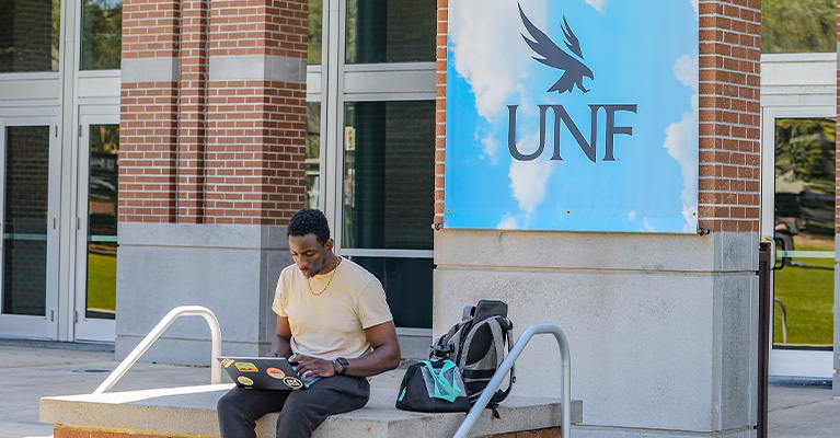 Student studying on a laptop in front of a UNF sign