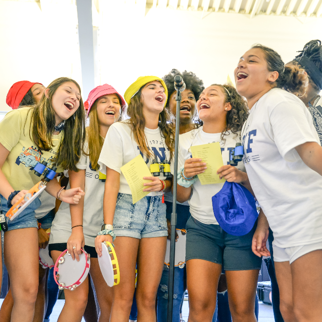 Group of female students in UNF t-shirts all around a standing microphone singing into it