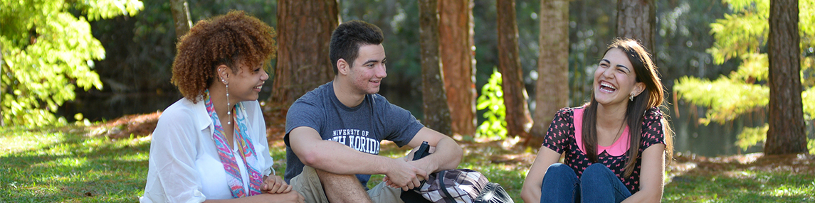 Three students talking to each other while sitting on grass near a campus lake