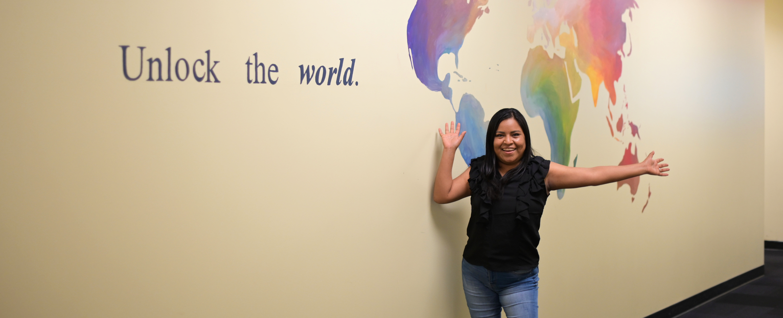 Female student holding her arms up and smiling in front of a map mural with text Unlock the World
