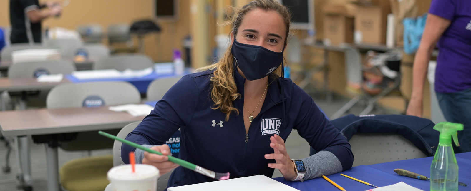 Female student wearing a face mask and UNF jacket sitting in a classroom holding a paint brush