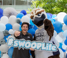 Male student holding a Swoop Life sign and pointing at the camera with Ozzie and balloon backdrop
