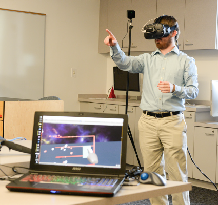 A male wearing a virtual reality headset and pointing into the air with a laptop showing what he's seeing