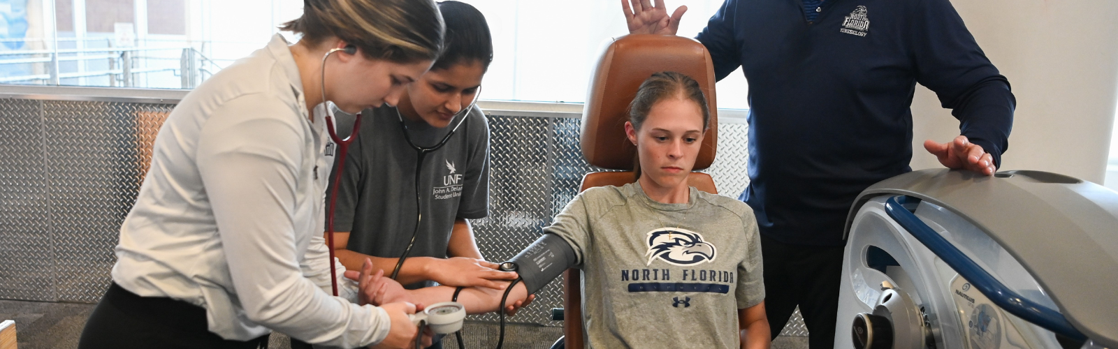 Two students with a stethescope and one student with a blood pressure cuff sitting in a chair. 