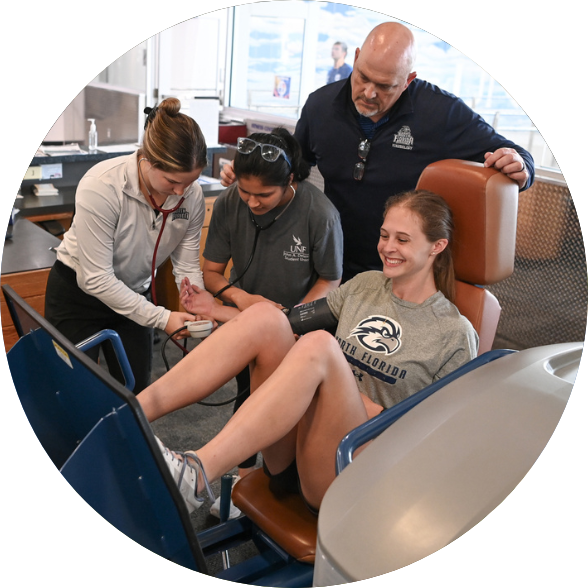 Faculty observing two students taking blood pressure from another student who is sitting on a leg press machine