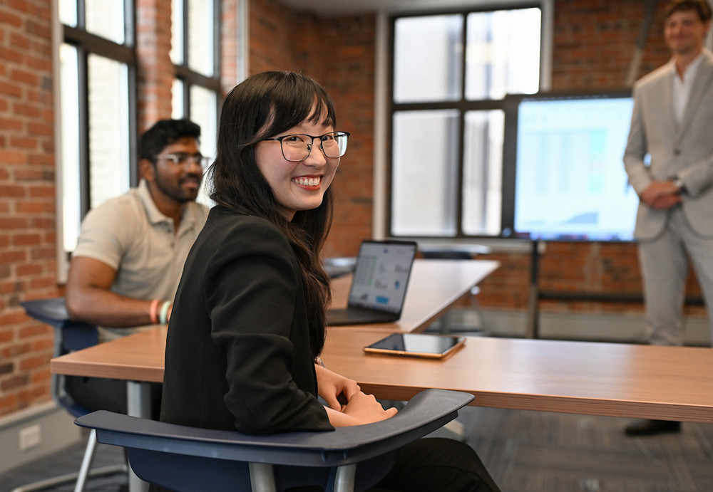 a student smiling at a desk with two other students during a presentation