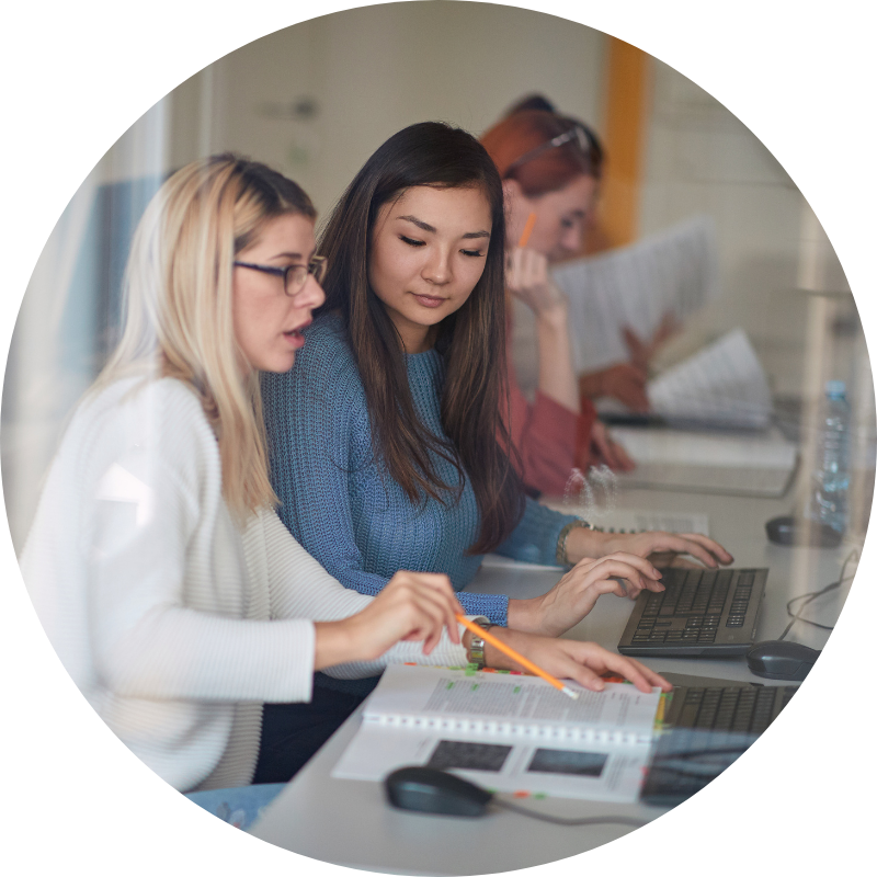 two girls at a computer reading from a notebook holding a pencil