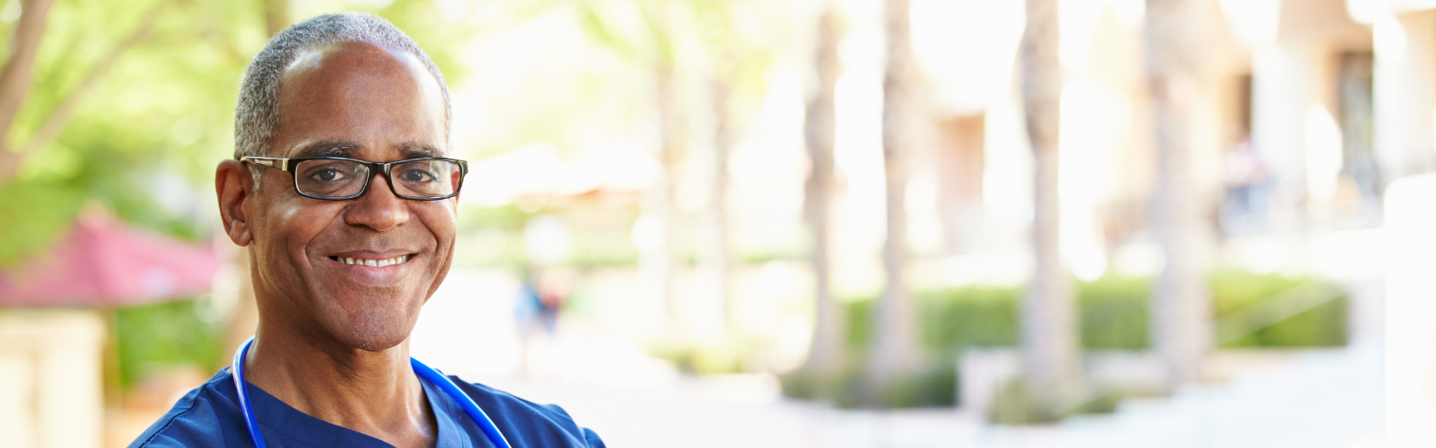 african american male nurse wearing black glasses and smiling, trees in the background