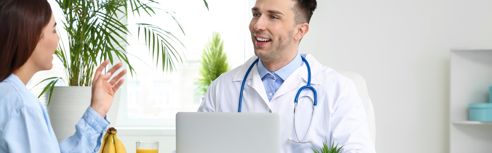 Man wearing a white coat and stethoscope talking to a woman at a desk