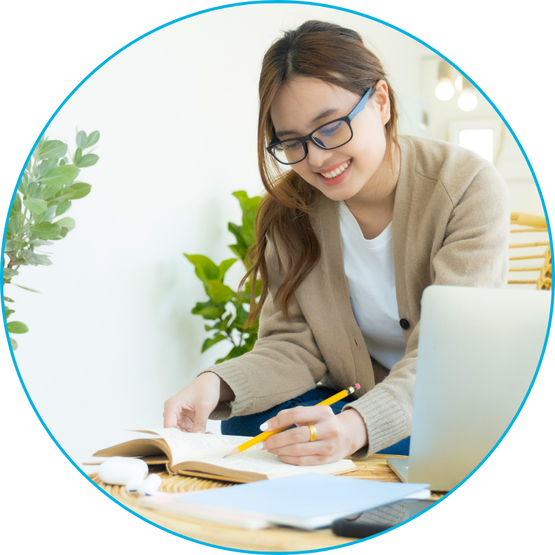 a woman wearing glasses, reading a book, holding a pencil, and sitting at a desk next to her laptop