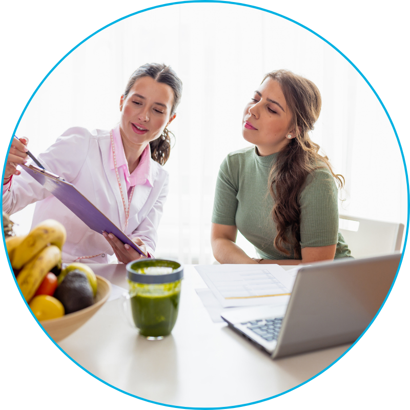 Two females, one dietitian, one dietetic student siting at a table with a clipboard, computer, papers, a cup of green juice and a bowl of fruit.