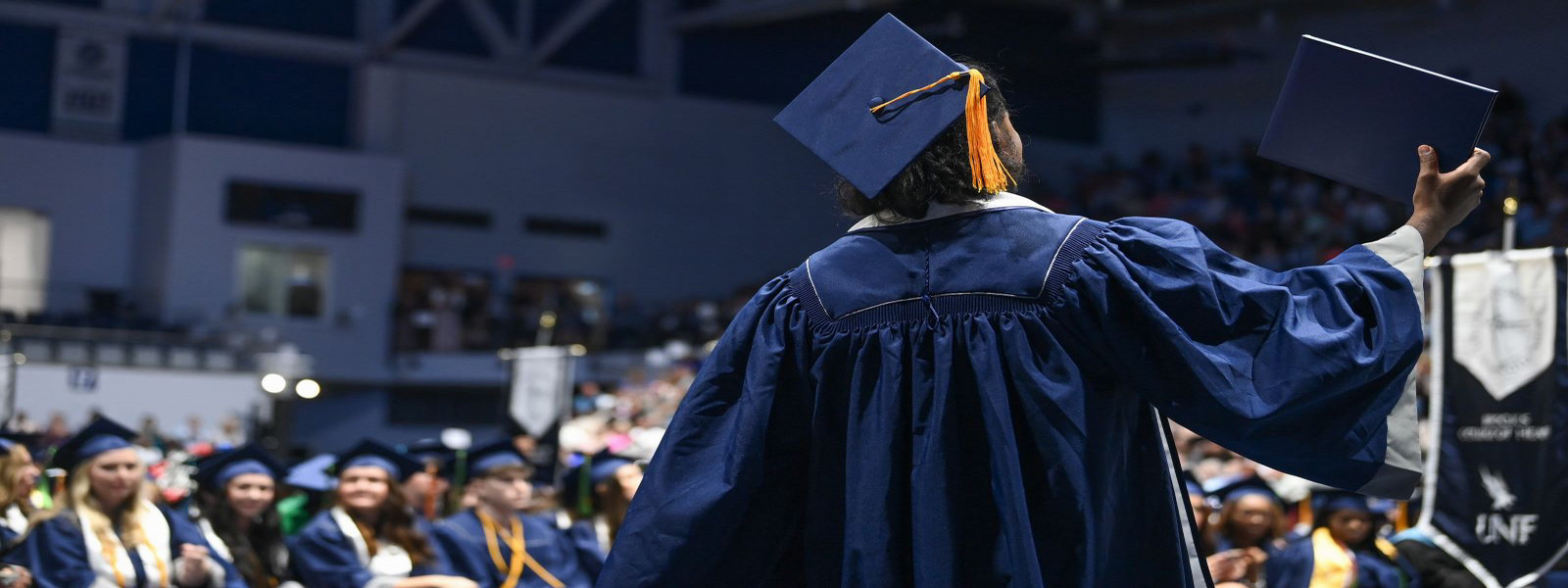 The back of a single student standing in his cap and gown in the crowd at graduation holding the diploma case