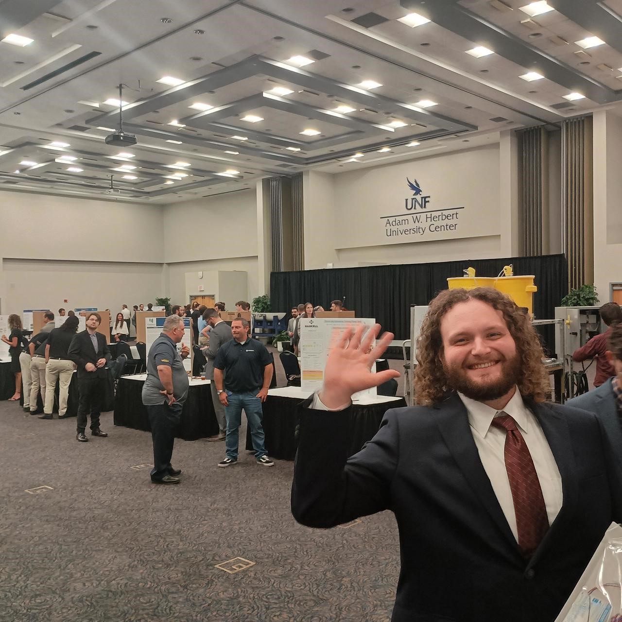 Student in suite at Innovation Day waving to the camera with people behind him in front of displays on tables talking among themselves.