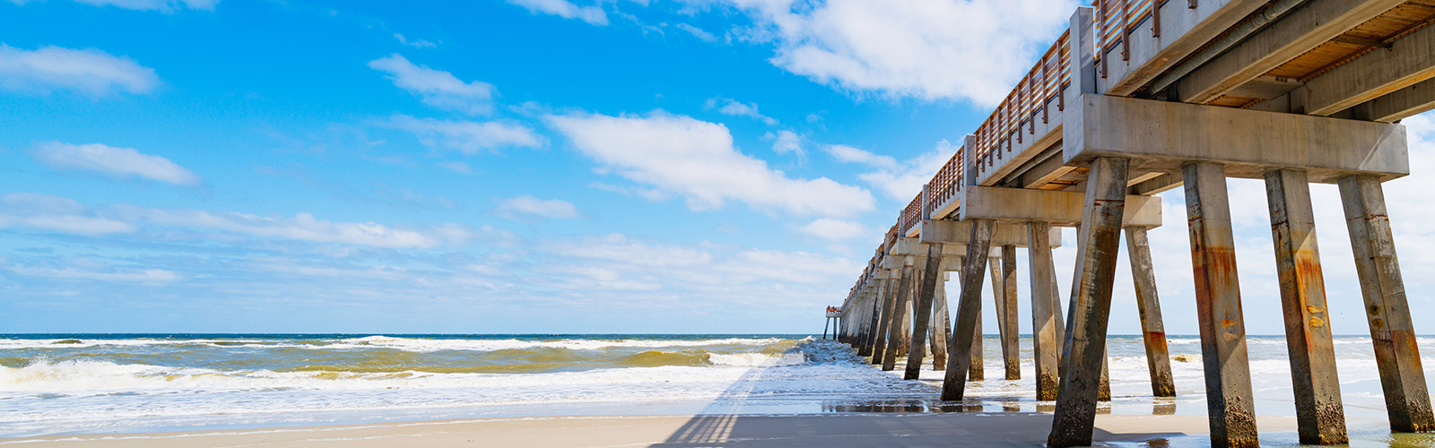 Seascape with sunny white sand beach, cloudy blue sky, shadows, and JAX Beach Fishing Pier in Jacksonville, North Florida