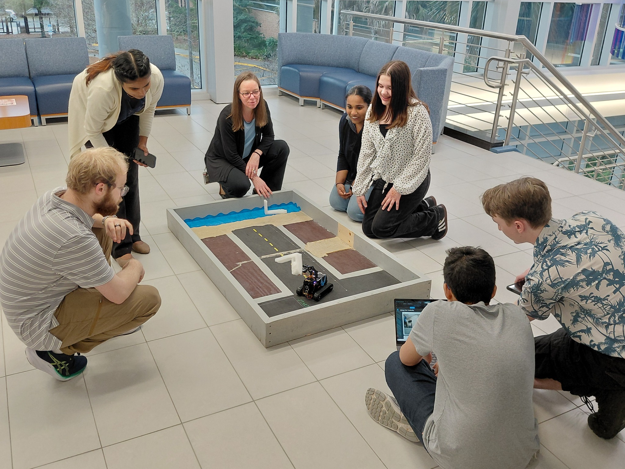 Four students, two instructors and one onlooker navigating a robot with tracks through and obstacle course. 