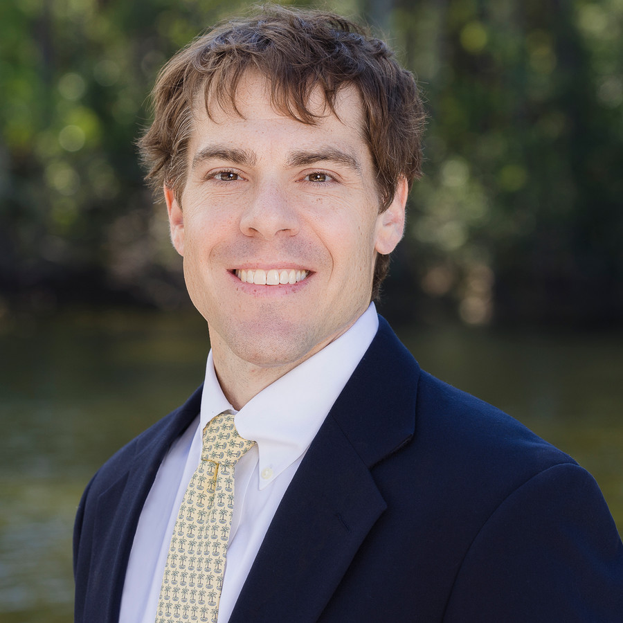 Headshot of Dr. Crowley in a Navy-blue suit jacket, white shirt and pale-yellow tie with light-blue palm trees printed symmetrically throughout the tie.